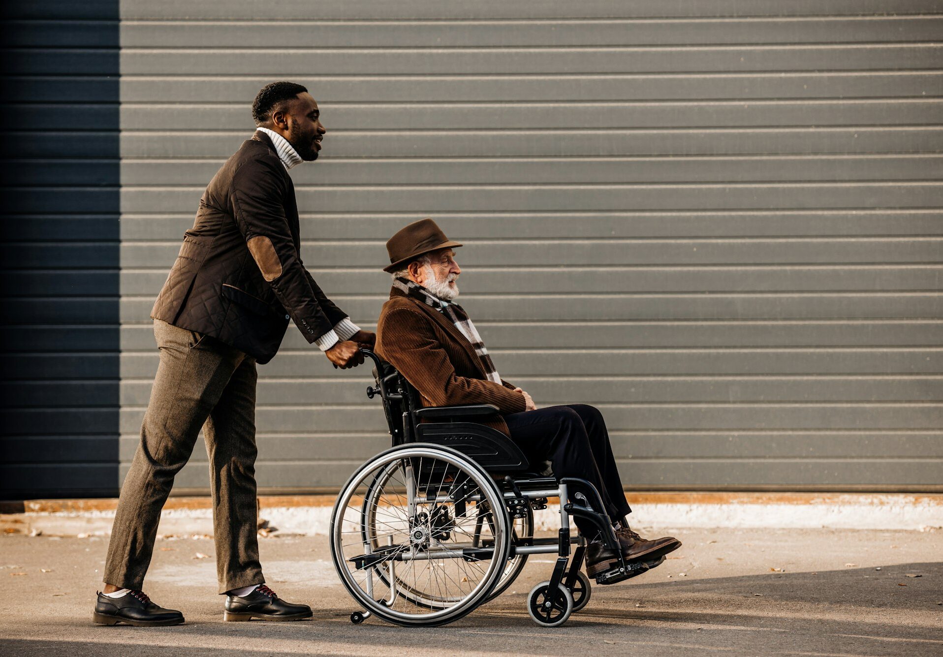 side view of senior disabled man in wheelchair and african american man riding by street